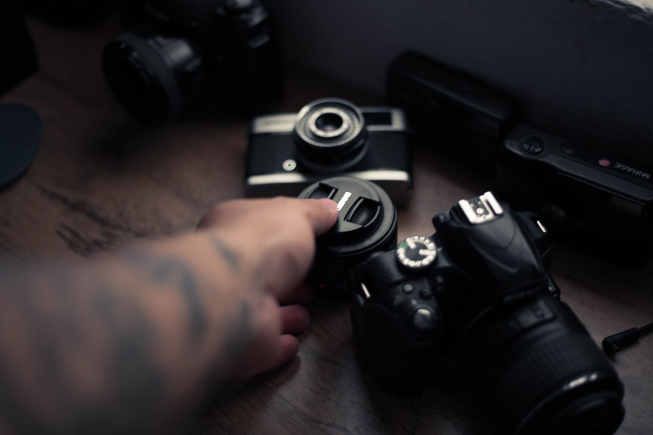 A person's hand holding a camera lens surrounded by various photography equipment on a wooden table.